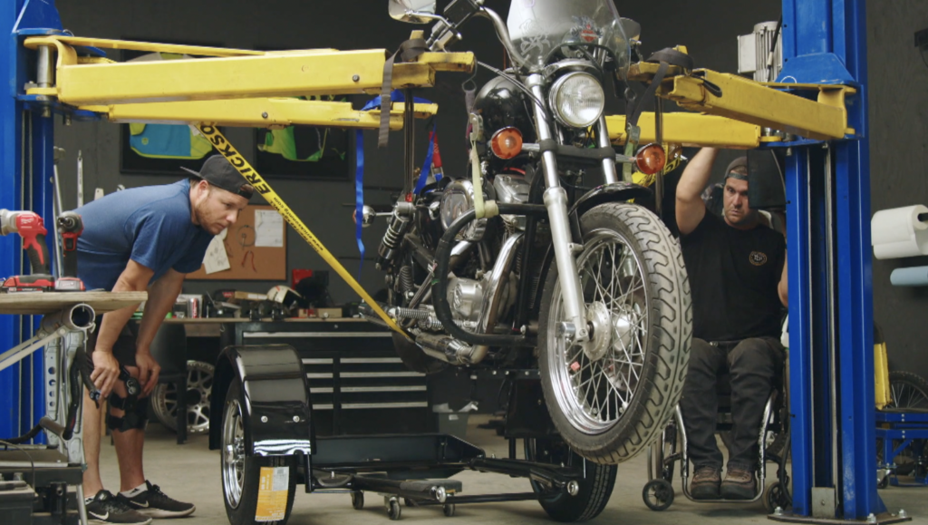 Tom and Bruce work on converting Roxie's motorcycle into a trike. The motorcycle is on a hoist. 